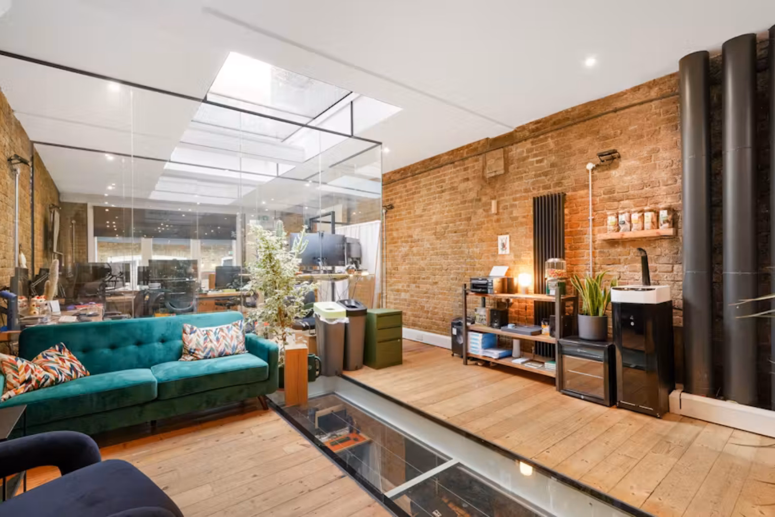 A green velvet sofa with patterned cushions sits on a wooden floor next to a glass floor panel and a raised wooden platform in an office space with exposed brick walls.