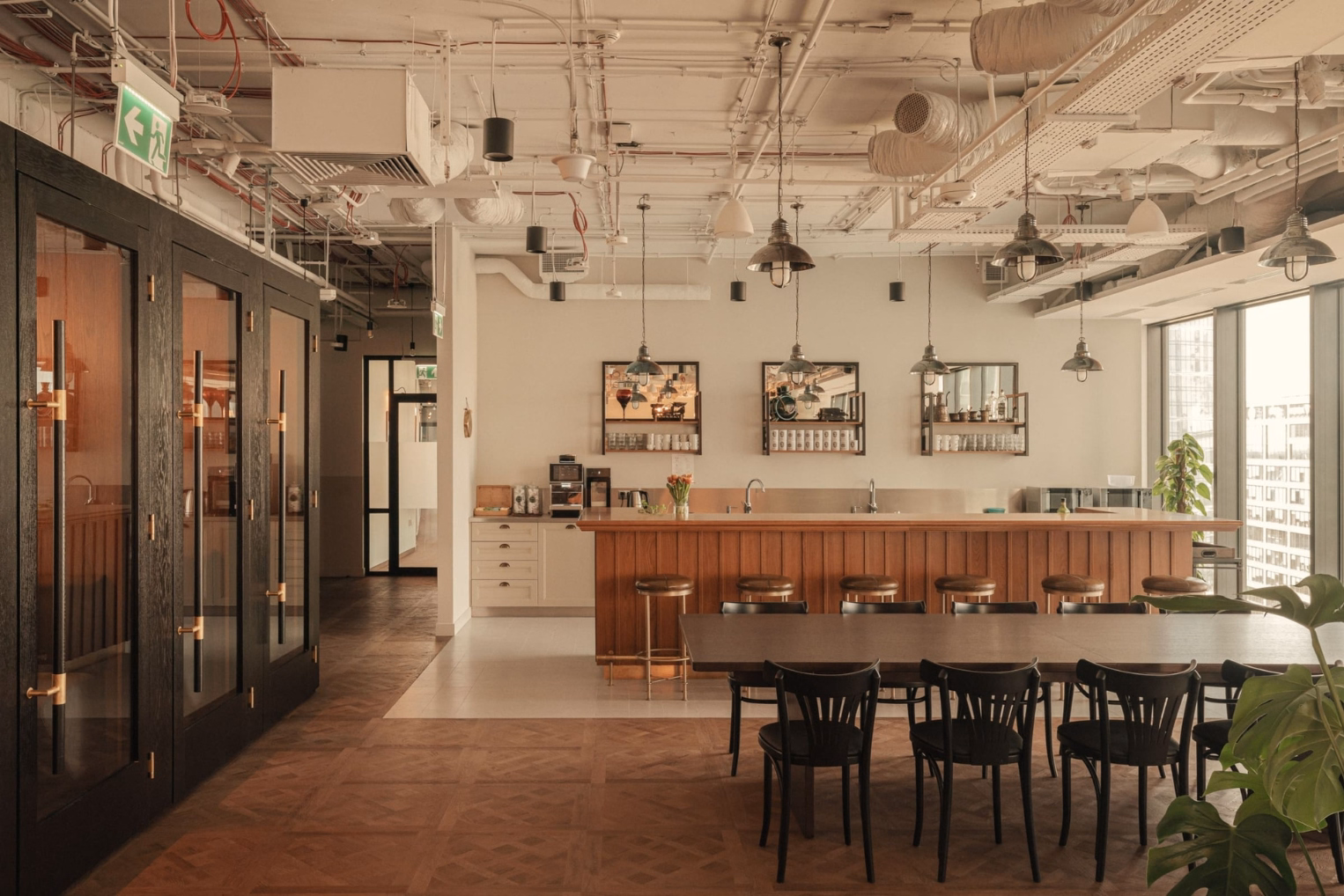 Black chairs surround a long dining table across from a wooden bar counter with stools in an open-plan office kitchen with exposed ceiling pipes.