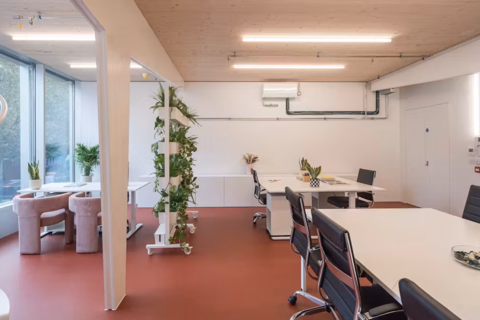 White desks with black rolling chairs sit on a reddish-brown floor next to a tall vertical plant stand in a modern workspace.