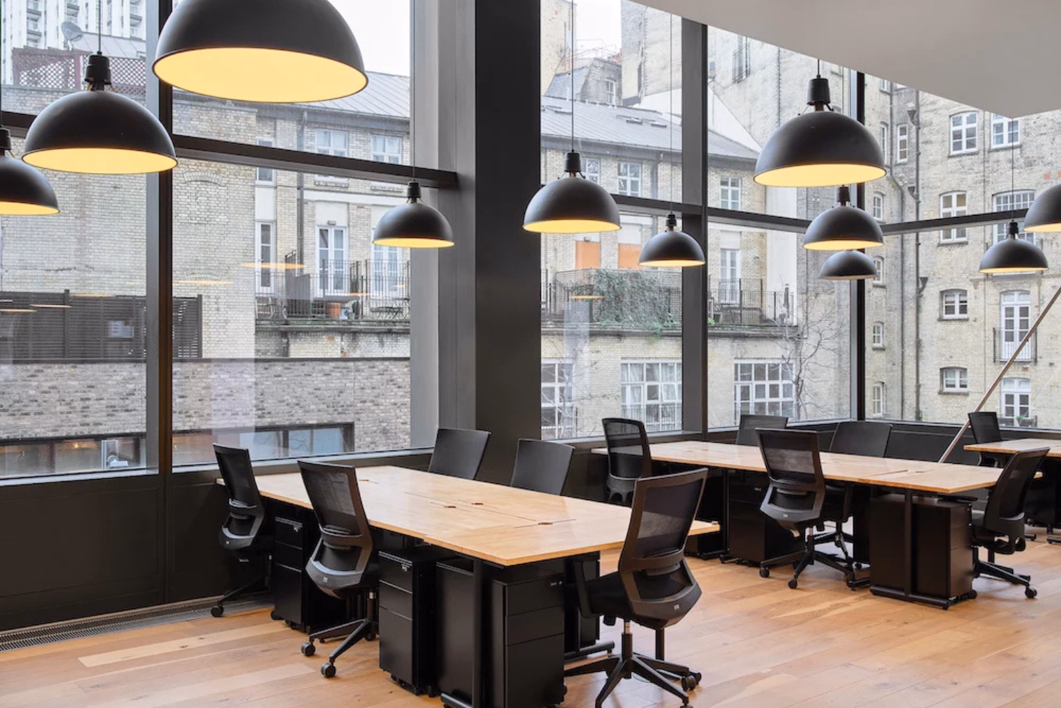 Black mesh office chairs are paired with light wood desks under dome pendant lighting next to floor-to-ceiling windows overlooking nearby buildings.