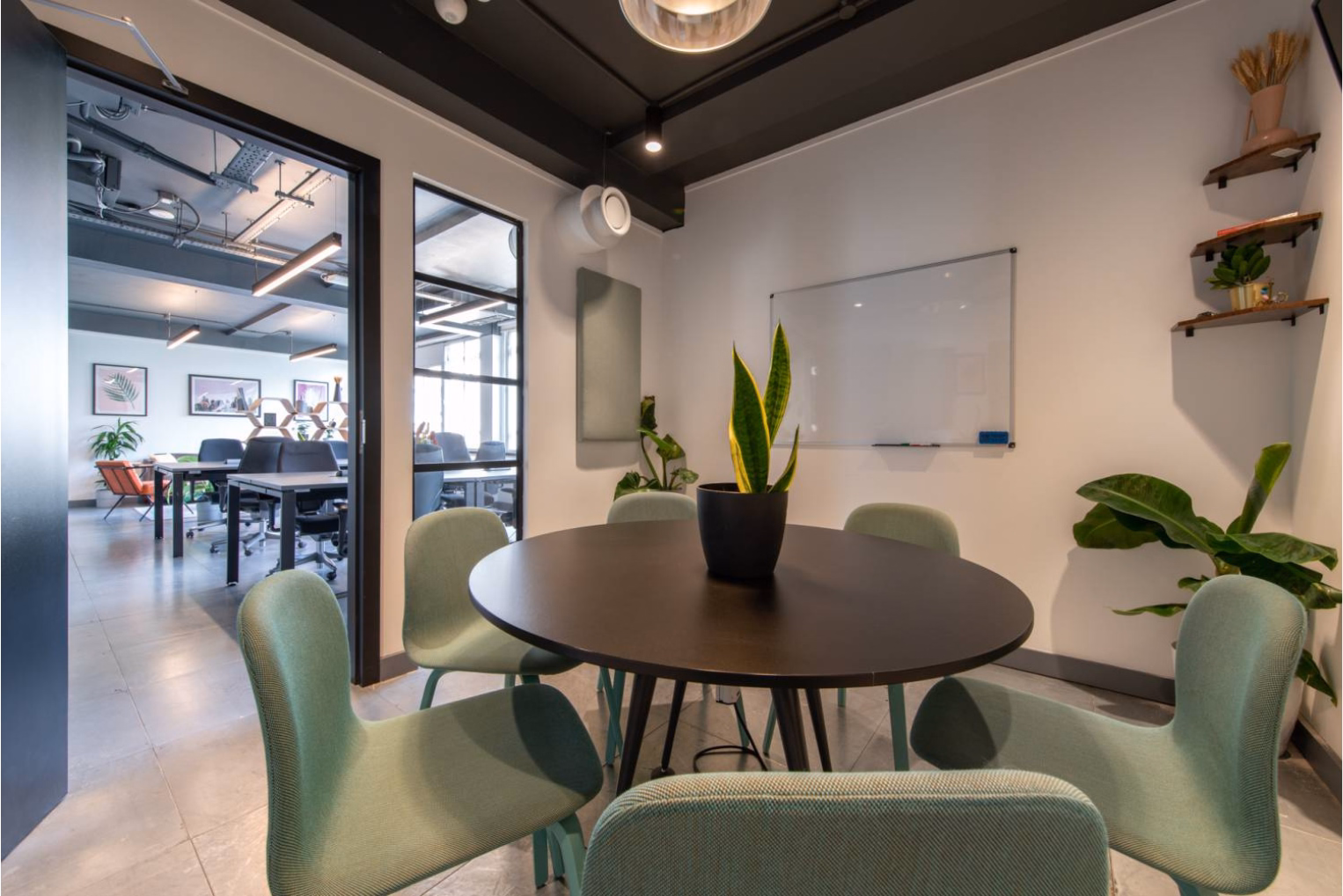 Light green upholstered chairs surround a round, dark wood table with a plant centrepiece in a meeting room with a whiteboard.