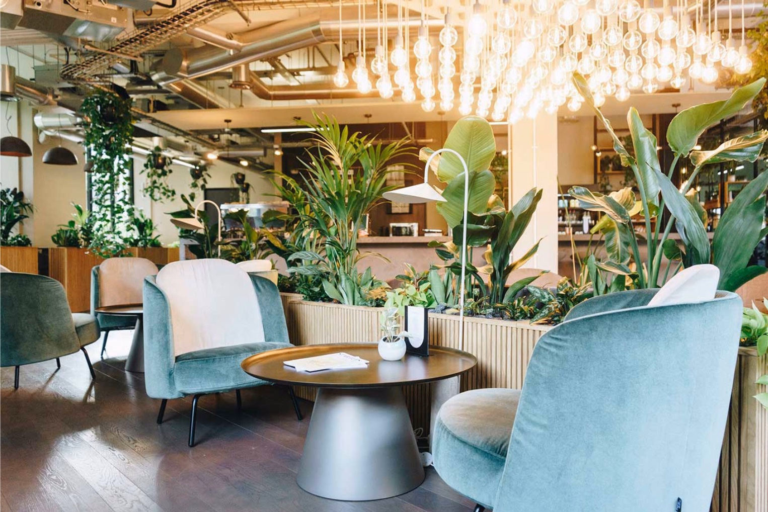 Green velvet armchairs are arranged around low coffee tables, framed by wood slatted planters filled with greenery and a large, glowing lightbulb installation above.