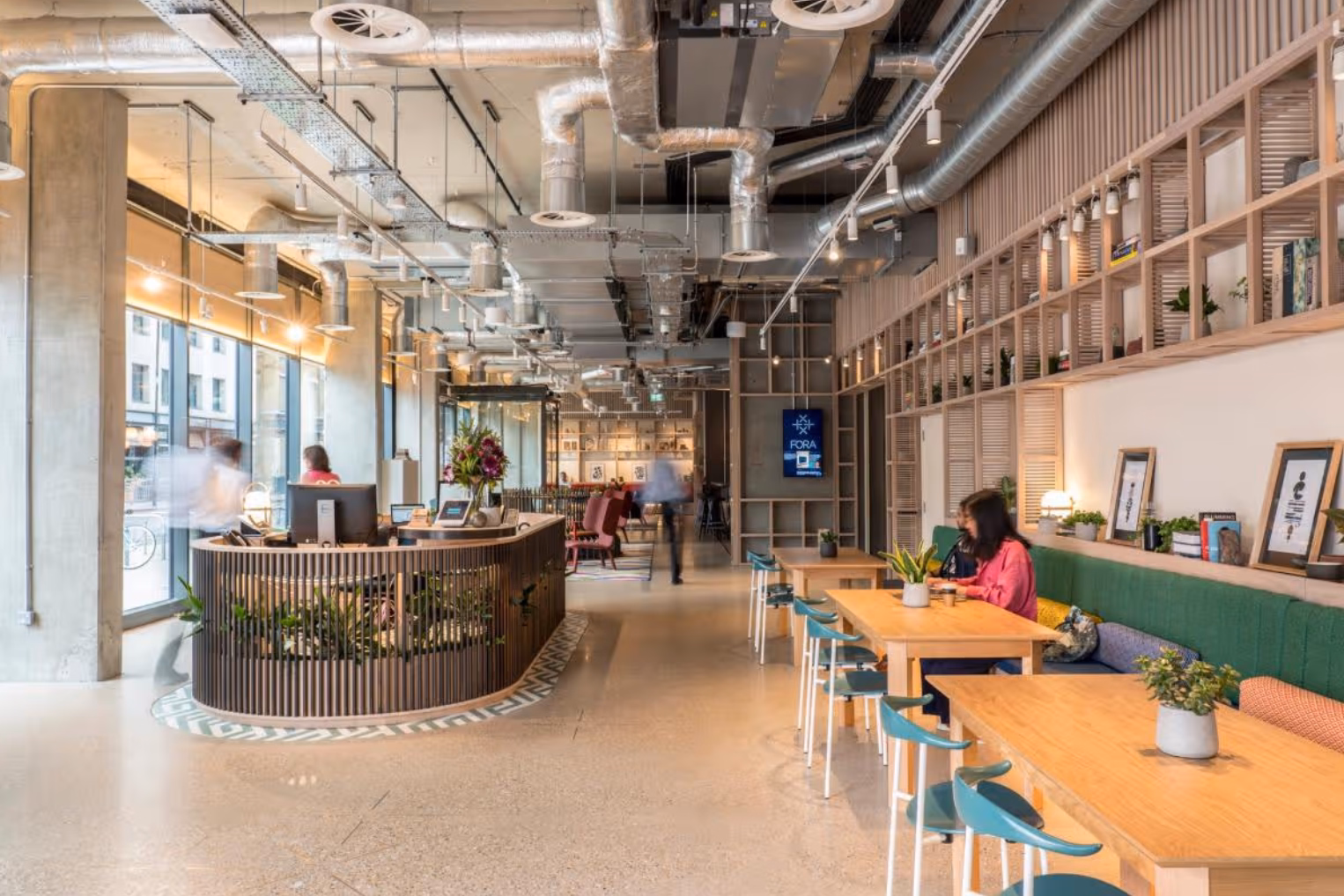 A curved, wood-slatted reception desk opposite wooden tables and a long banquette seat, beneath a ceiling with exposed ductwork.