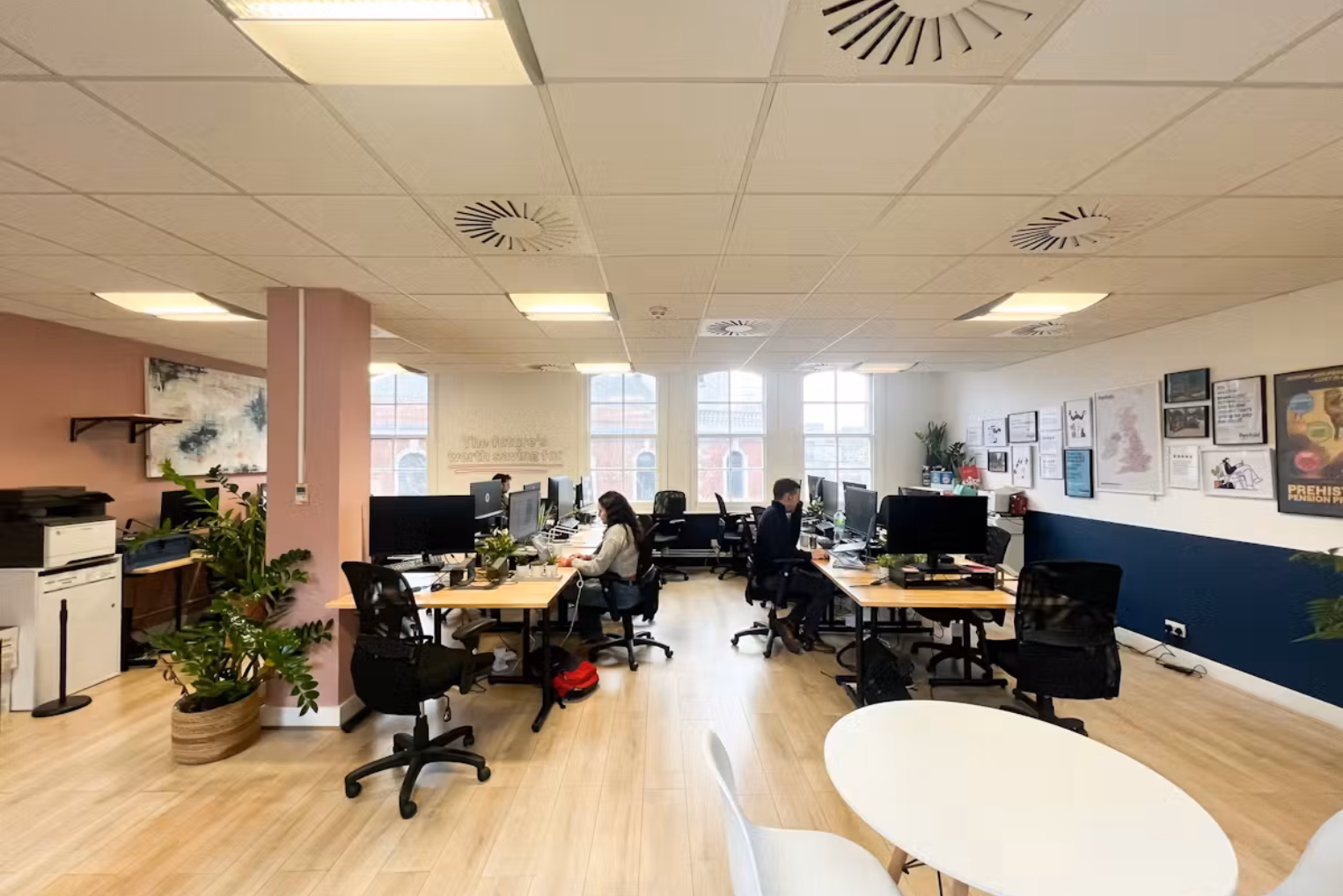 An open-plan office space with rows of wooden desks with computers, black mesh chairs, pink walls, and large windows that fill the room with natural light.