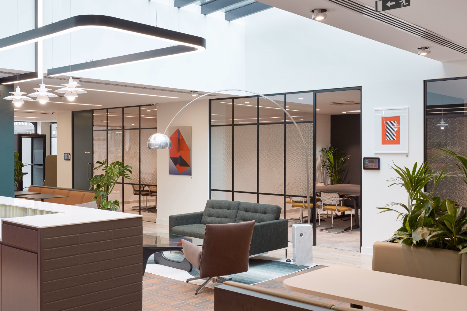 Open-plan lounge and reception area with a dark green sofa and a leather armchair beneath a large, arched metal floor lamp, with black-framed glass partitions.