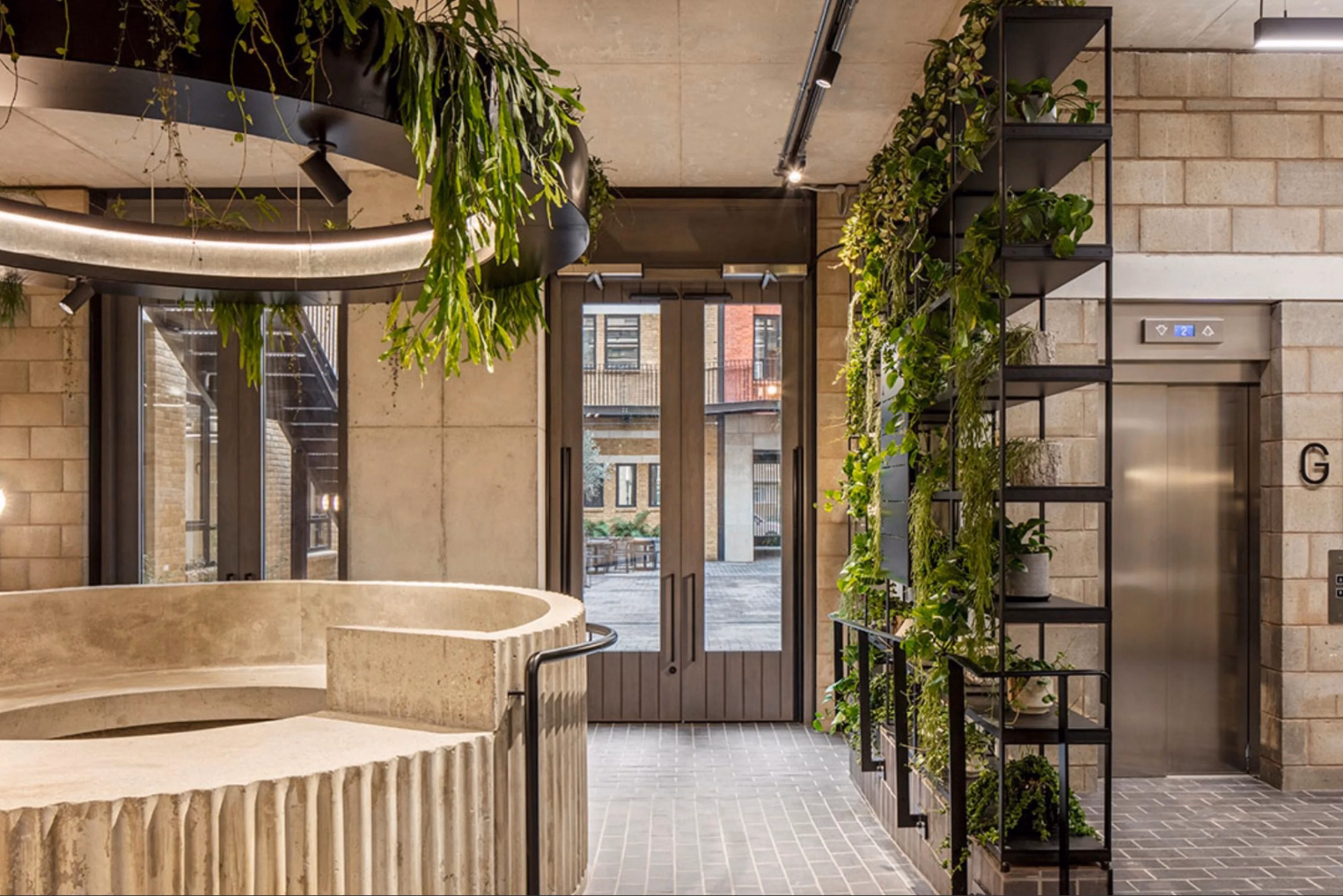 Entrance area with a curved, concrete desk, a large circular overhead planter, and a tall metal shelving unit with trailing green plants next to an elevator.