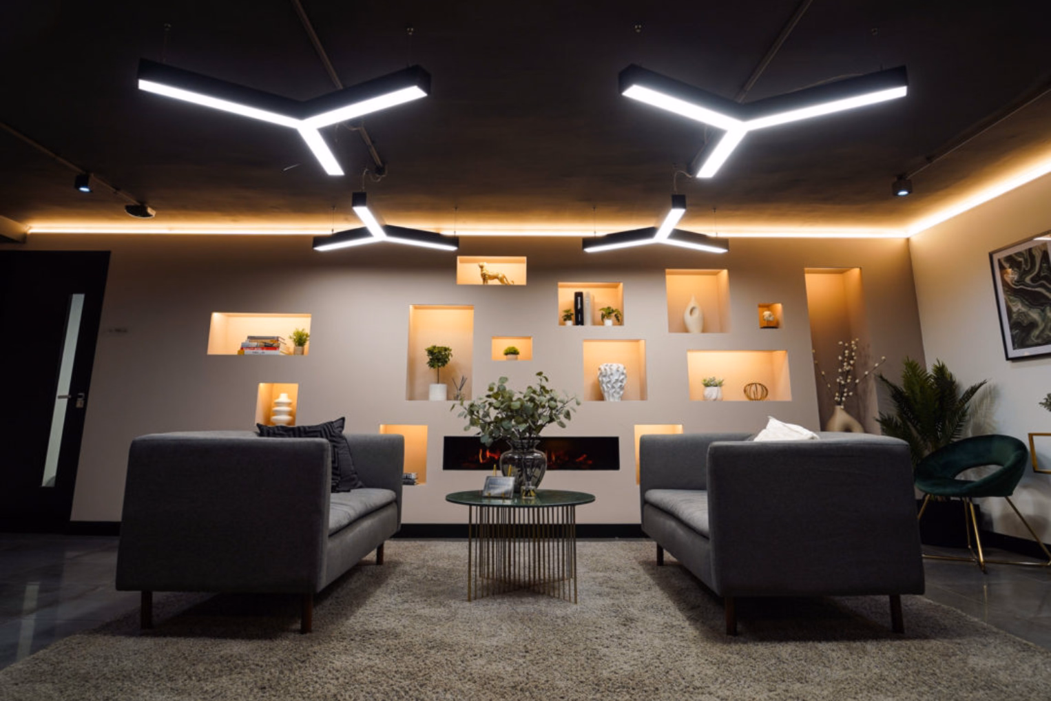 Two grey sofas face each other over a coffee table in a lounge area, with overhead lights and a back wall with an illuminated shelving display.