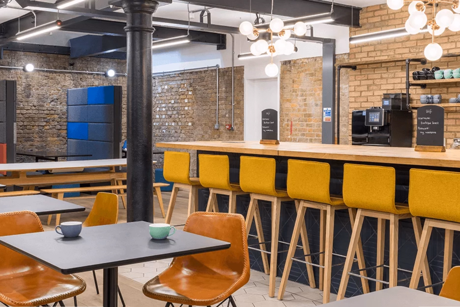 An industrial-style office kitchen and breakout space with a wooden breakfast bar lined with yellow stools, exposed brick walls, a black pillar, and globe lighting.