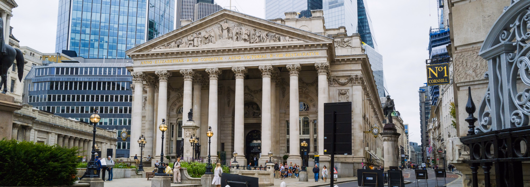 A stone building with massive columns and an intricate sculpture, contrasted by towering modern blue-glass skyscrapers directly behind it.