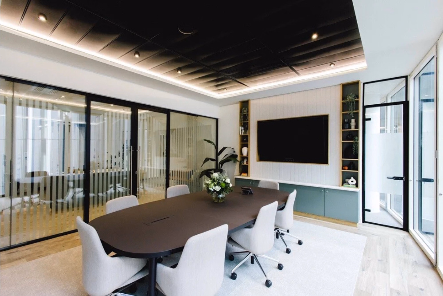 A dark brown table surrounded by light grey chairs on a white rug with a white panelled accent wall with sage green cabinets and a black slatted ceiling.