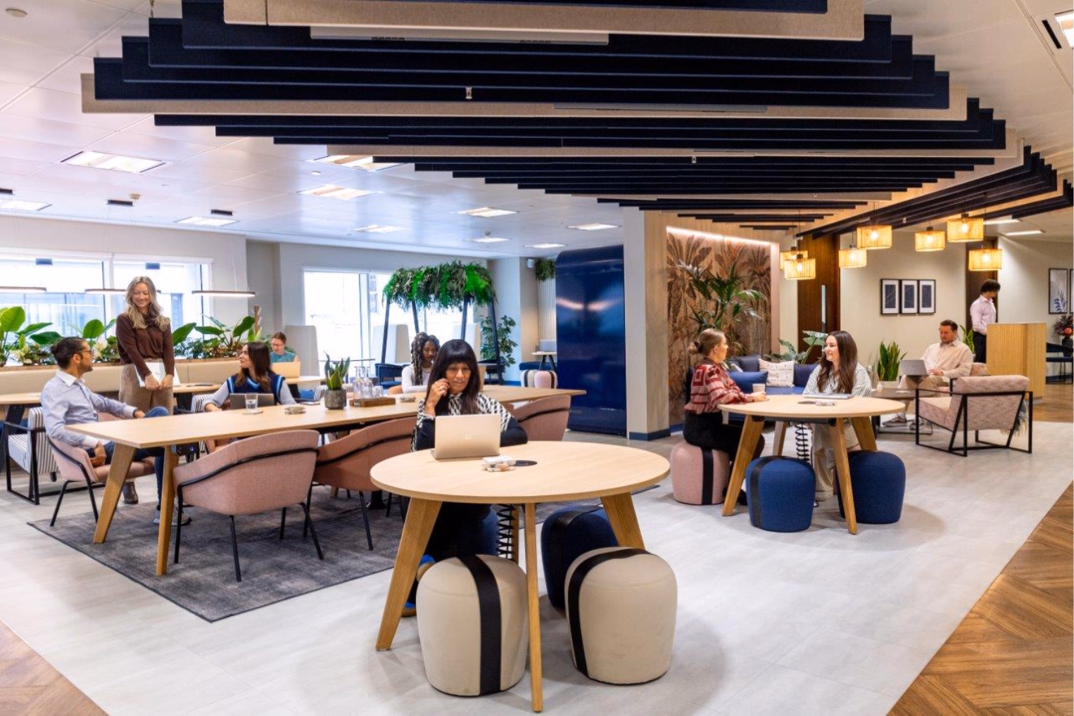 People sit and work at light wood tables with pink chairs and blue and beige pouffes in a lounge with a dark blue slatted ceiling installation.