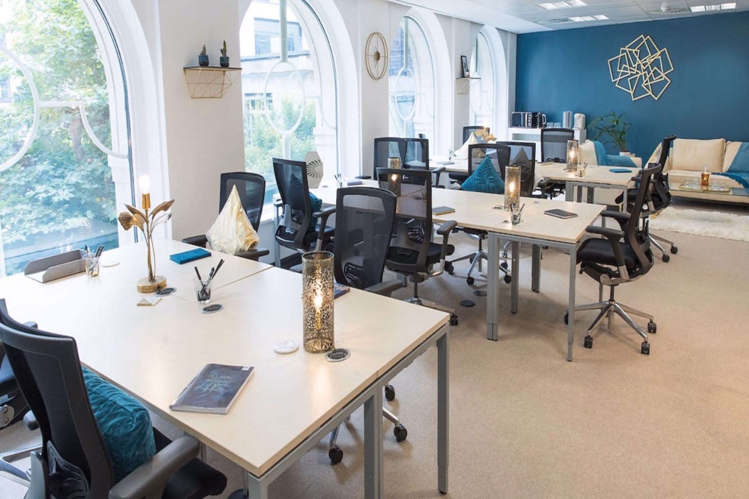  Rows of white desks with black mesh chairs next to arched windows in a bright office space with a deep blue accent wall and a cream-coloured sofa.