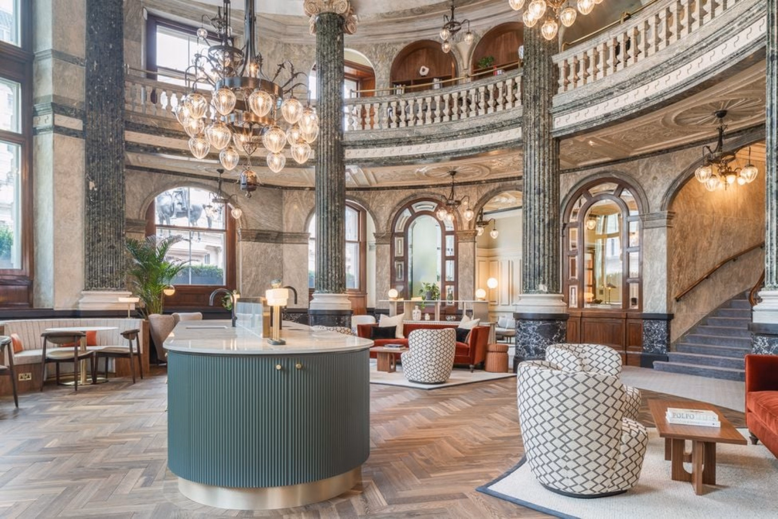 Patterned club chairs and red velvet sofas on a herringbone wood floor featuring green marble columns and glass chandeliers.