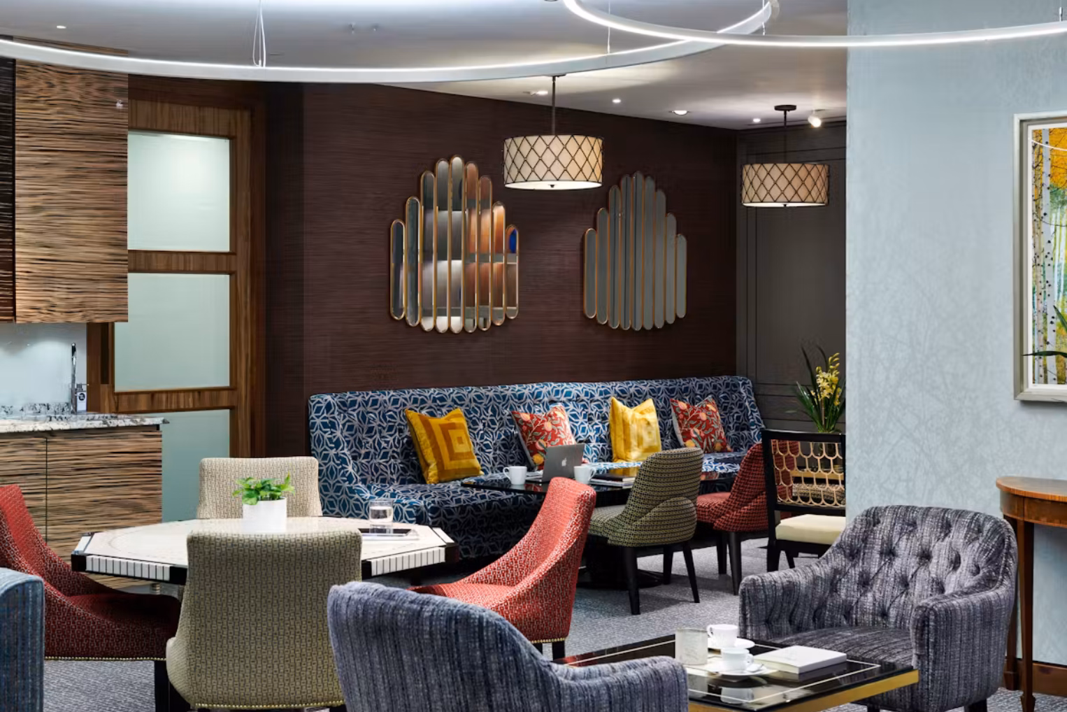 Colourful, patterned armchairs surround a white table across from a long blue patterned sofa with yellow and red cushions against a dark brown wall.
