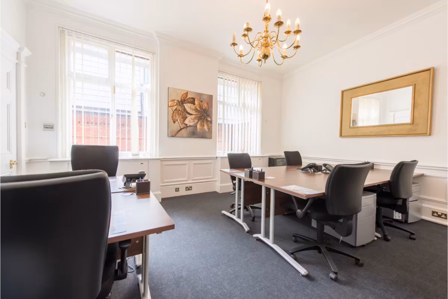 Black chairs around wooden desks on a dark grey carpeted floor in a white-walled office with a gold chandelier and a large gold-framed mirror.