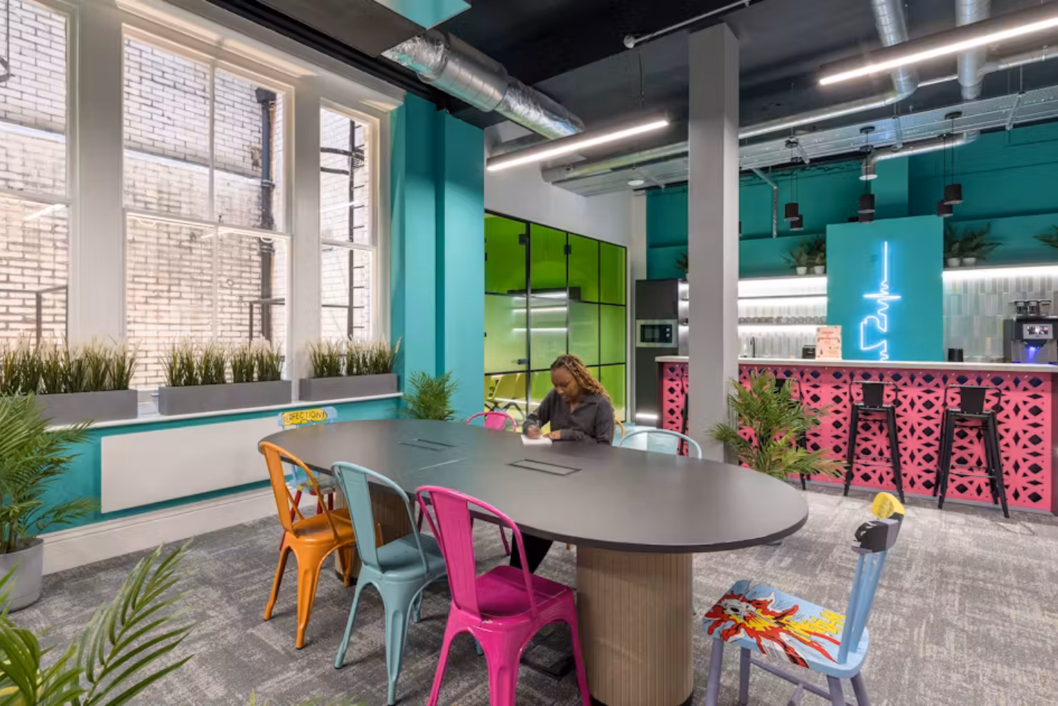A person sits writing at a black table surrounded by colourful chairs. A kitchen with turquoise walls, lime green glass partitions, and a pink patterned bar counter.