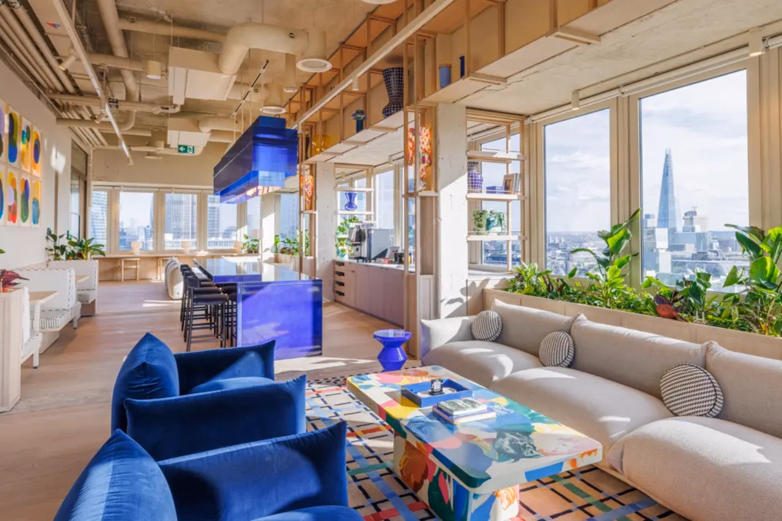 Bright blue velvet armchairs and a multi-coloured, patterned coffee table with a beige sofa and large windows that overlook a city skyline.