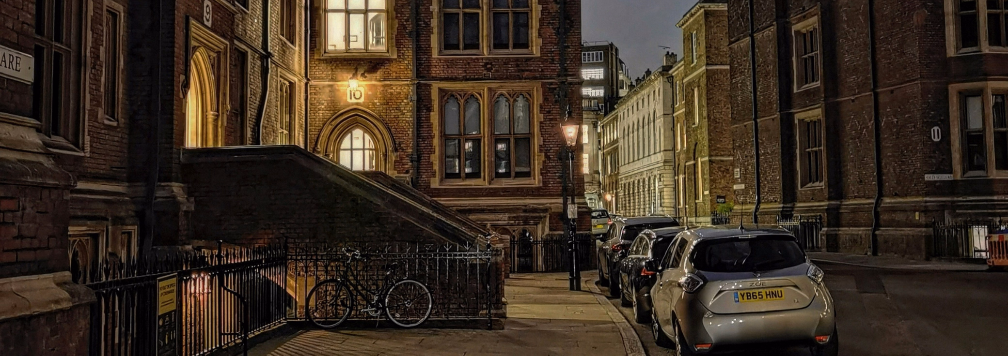 A silver hatchback is parked on a city street lined with dark brown brick buildings at night, with warm yellow light glowing from arched doorways and windows.