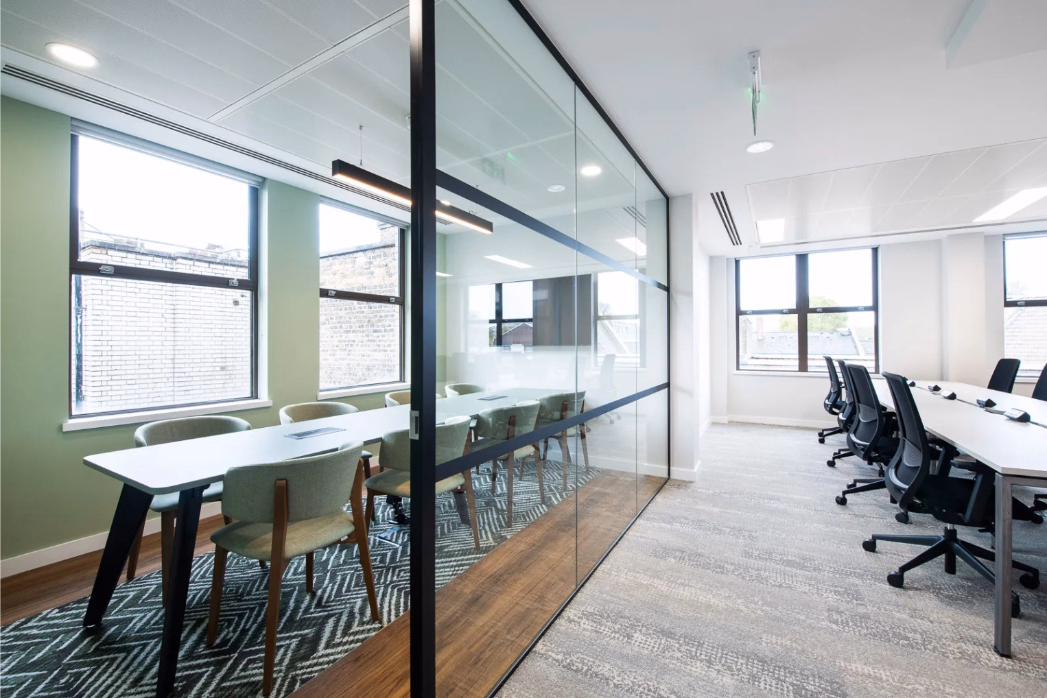 Green chairs surround a long white table inside a glass-partitioned meeting room, adjacent to an office with white desks and black mesh chairs on a grey carpeted floor.