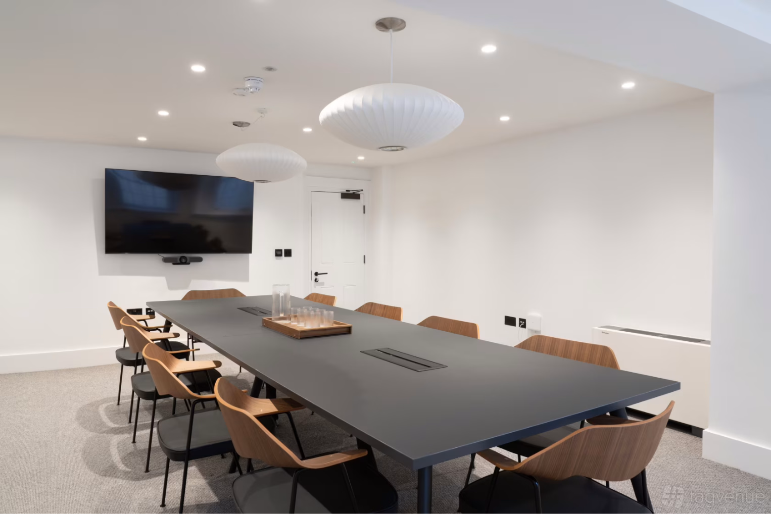  A dark grey table surrounded by brown wooden chairs with black seats sits on a grey carpeted floor with white walls and spherical lights.