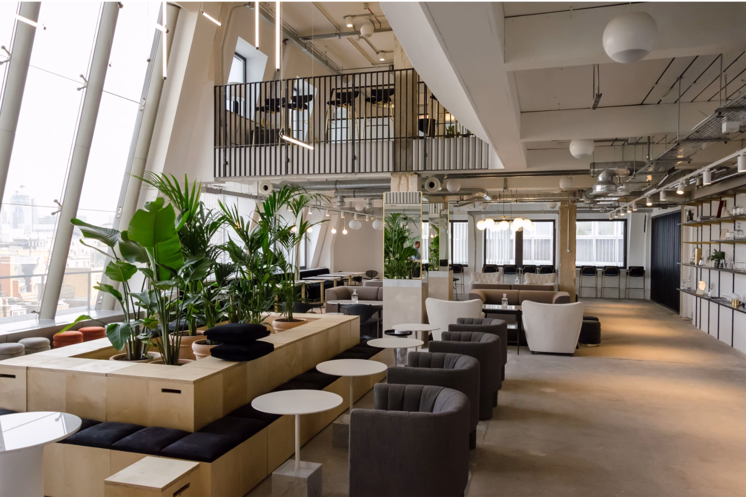 Grey armchairs and white pedestal tables next to a tiered wooden seating structure filled with large green plants.