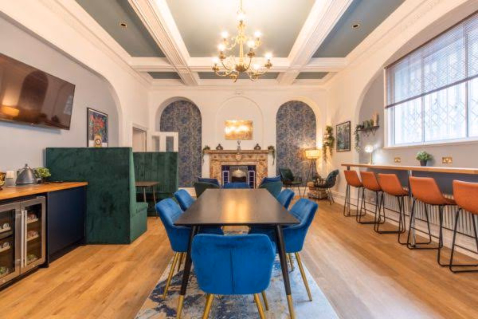 Blue velvet chairs surround a dark wooden table; there are green high-backed booths and a row of orange barstools.