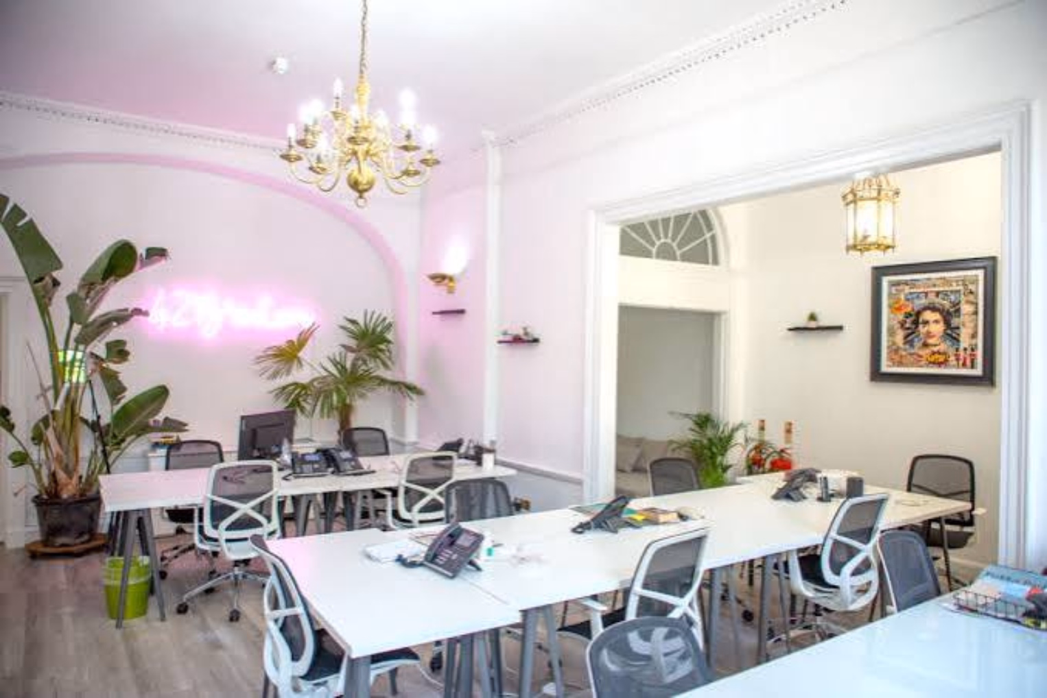 White desks with white-framed mesh chairs are across light grey wood floors in a room featuring a pink neon sign.