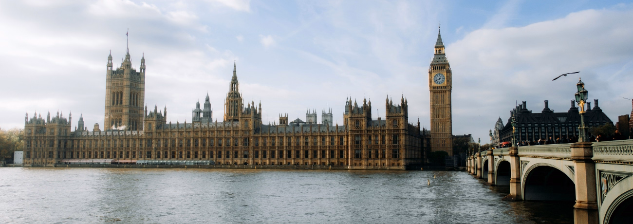 The Palace of Westminster and Big Ben along the grey River Thames next to Westminster Bridge, under a cloudy blue sky.