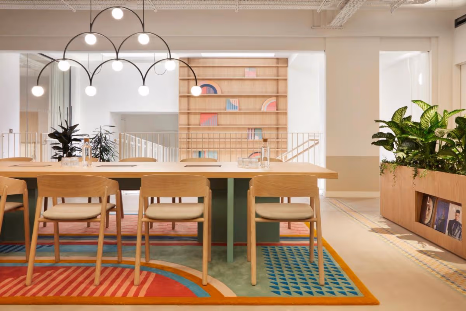 Light wood chairs around a long table on a multi-colored geometric rug beneath a black multi-bulb arched chandelier.