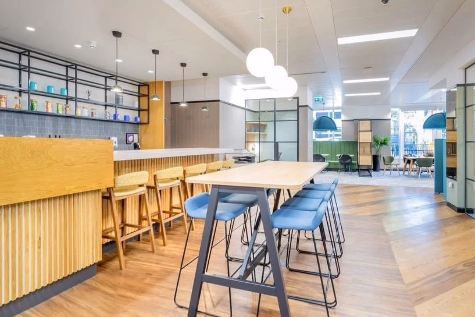 Blue barstools at a long, light wooden high-top table next to a fluted wooden bar counter with yellow stools under hanging white globe lights.