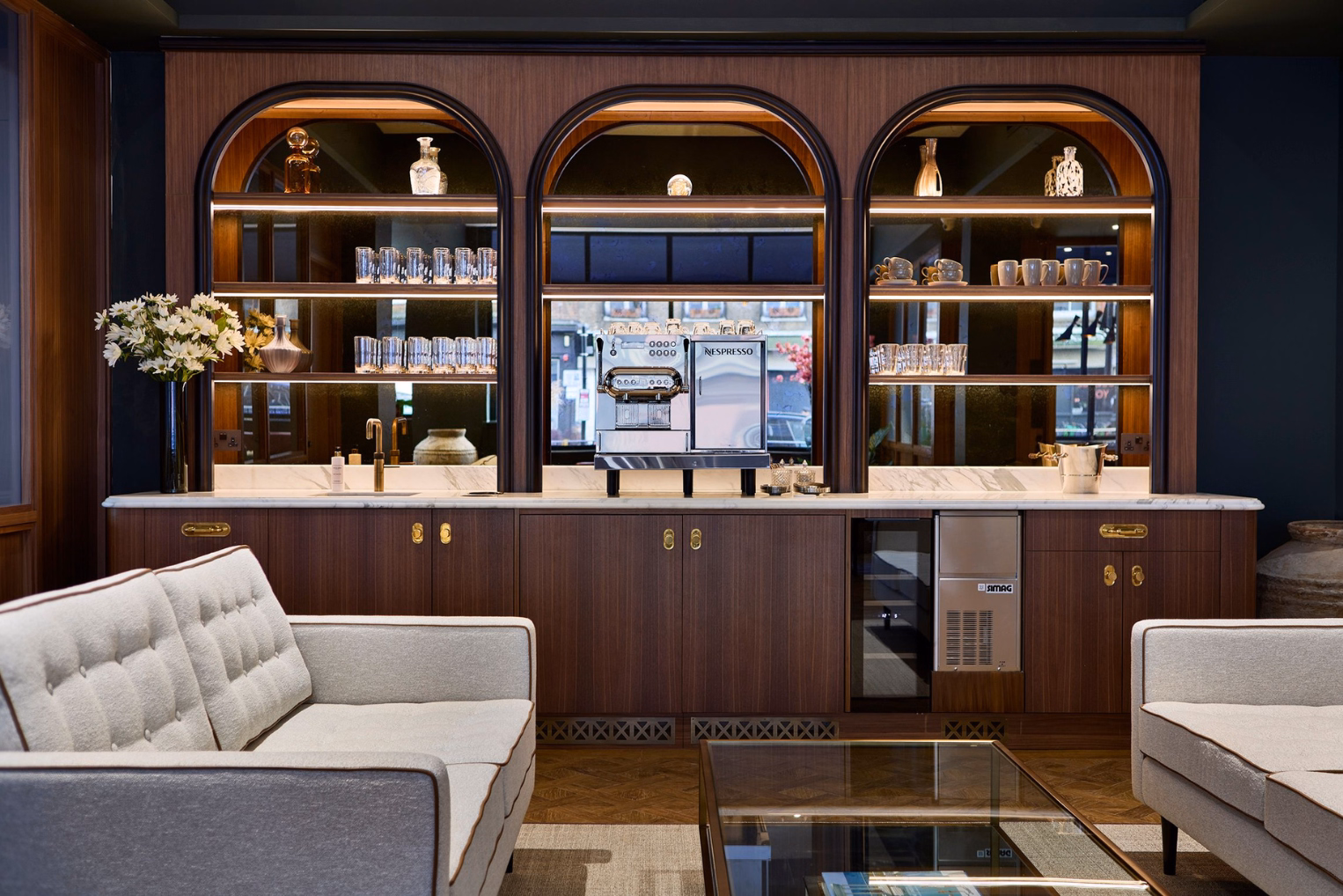 Grey sofas, a glass-topped coffee table, a dark wood beverage station featuring three lit, arched mirror-backed alcoves that hold glassware.
