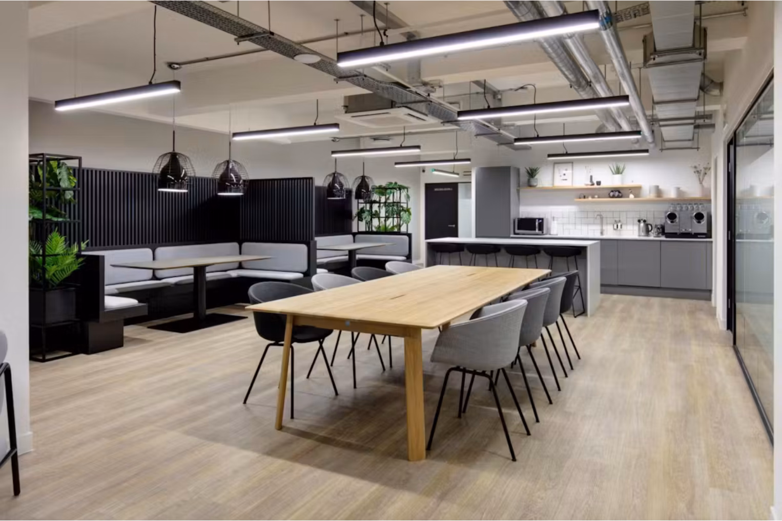 Grey and black chairs surround a long wooden table next to grey and black dining booths and a grey kitchen area.