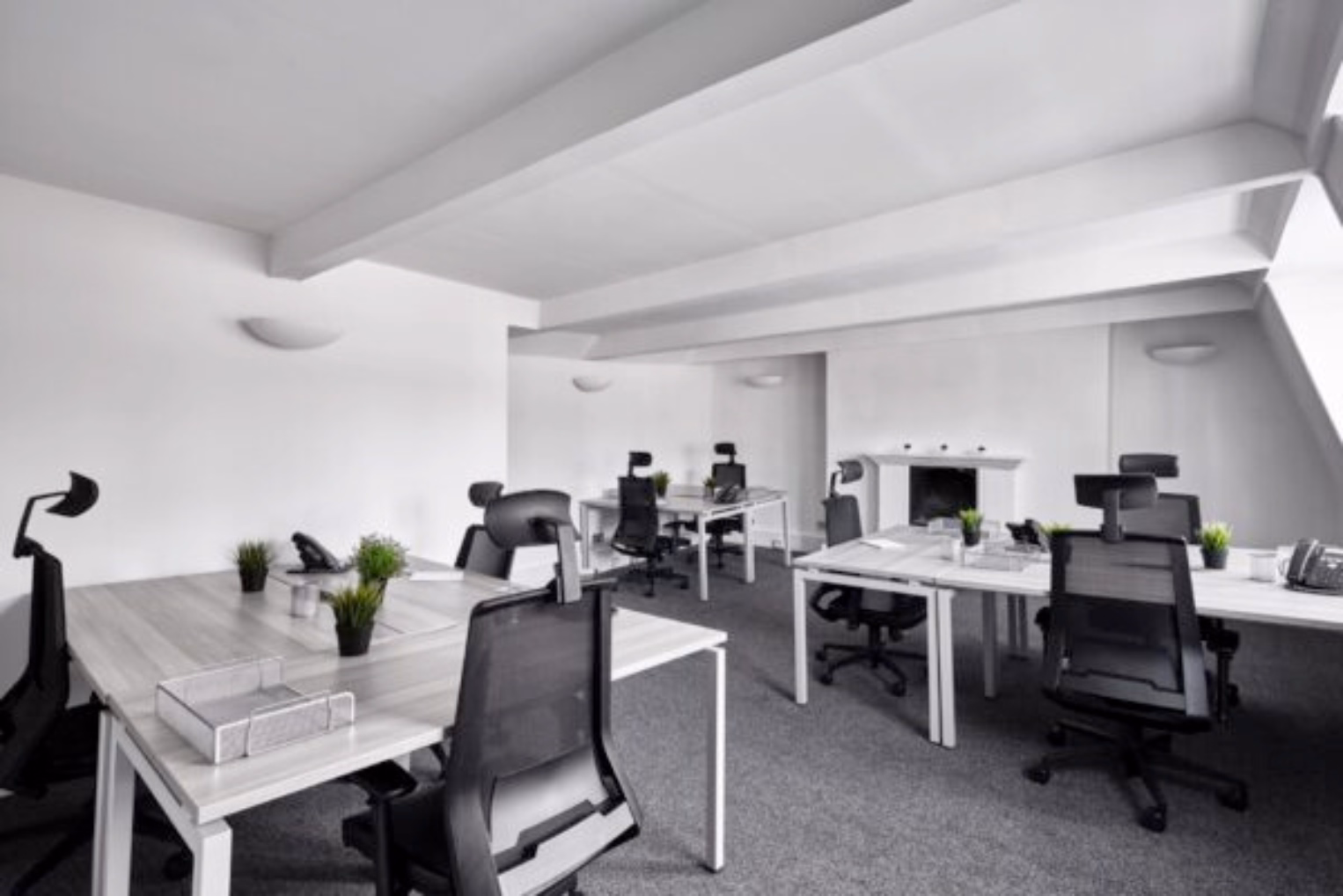Black chairs with high mesh backs at grey desks on a grey carpeted floor in a office with white walls and a sloped ceiling.