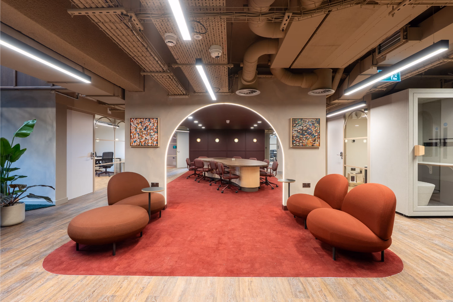 Orange rounded armchairs and a matching ottoman on a red carpet under an illuminated arched entryway leading to a conference table with dark red chairs.