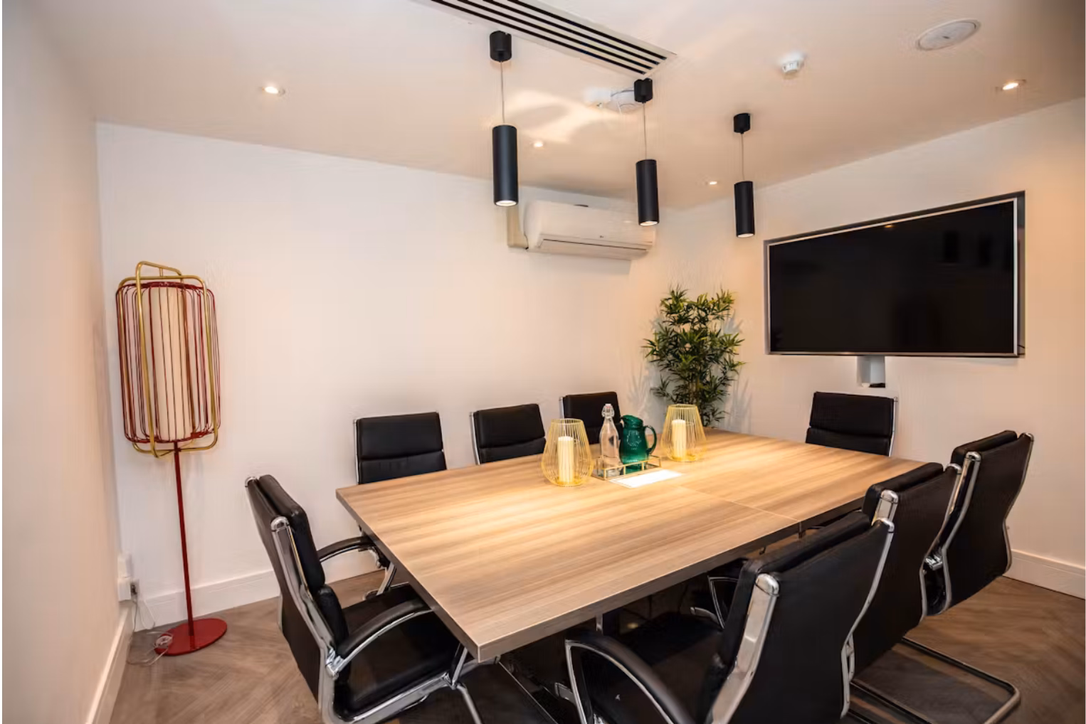 Black leather chairs surround a light brown wooden table in a white-walled room with black pendant lights and a wall-mounted TV.