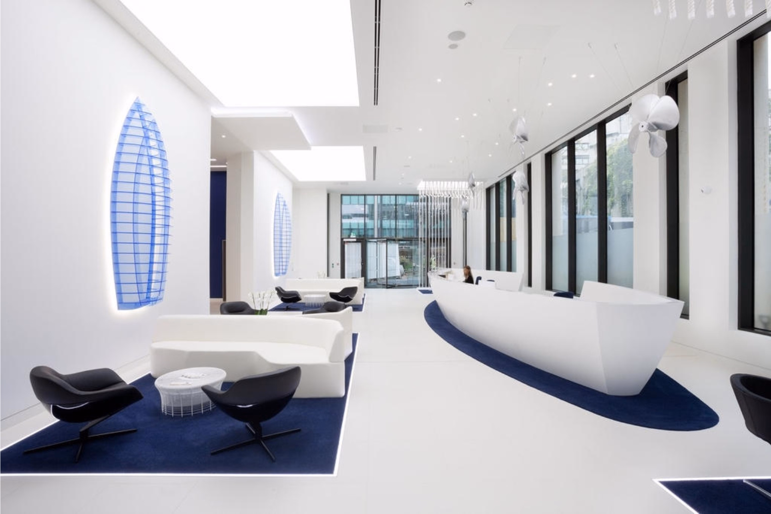 A curved white reception desk in a waiting area with white sofas and black swivel chairs on dark blue rugs.