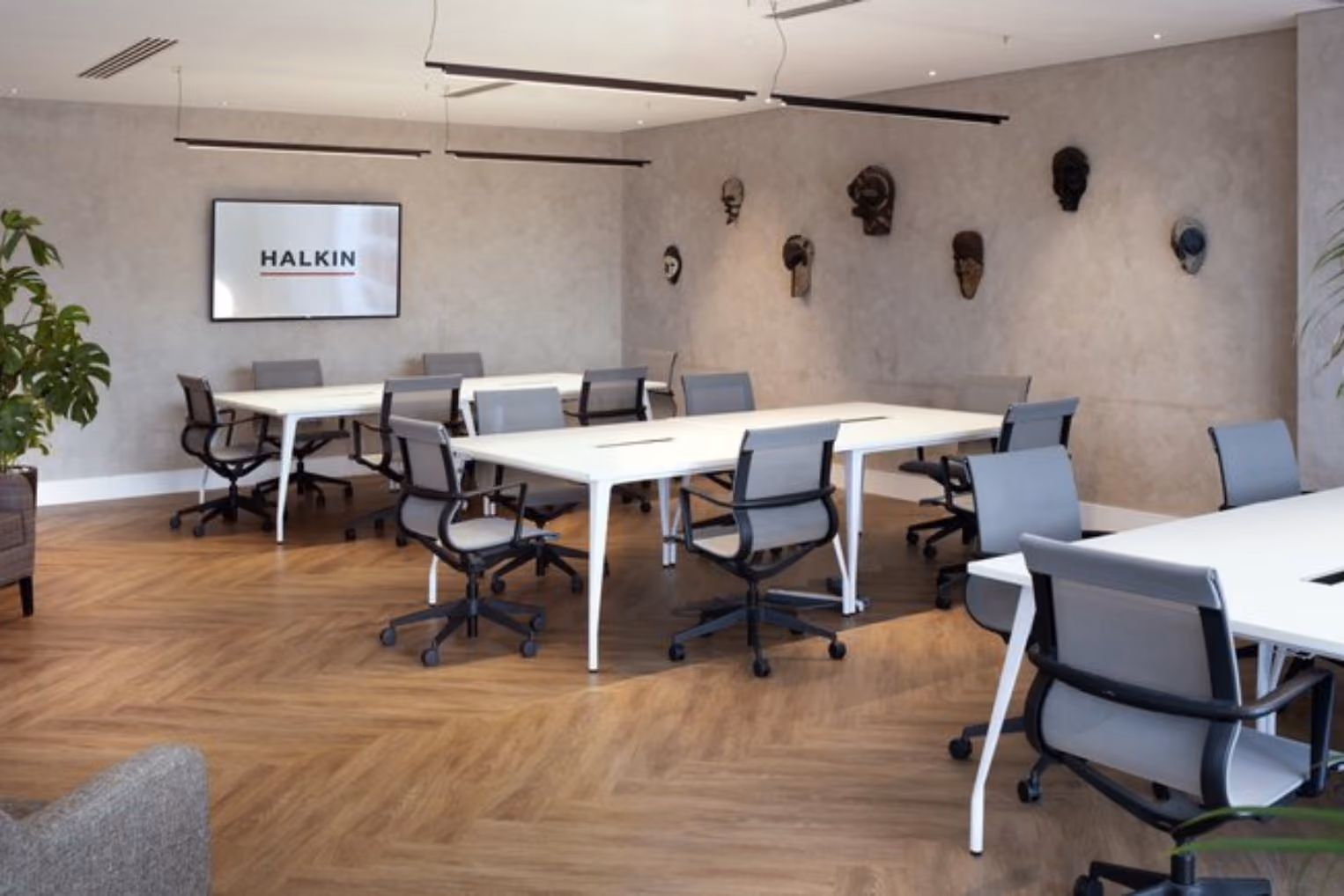 Grey rolling chairs with white desks on a wood floor, in a room with decorative masks on the walls and a TV displaying the "HALKIN" logo.