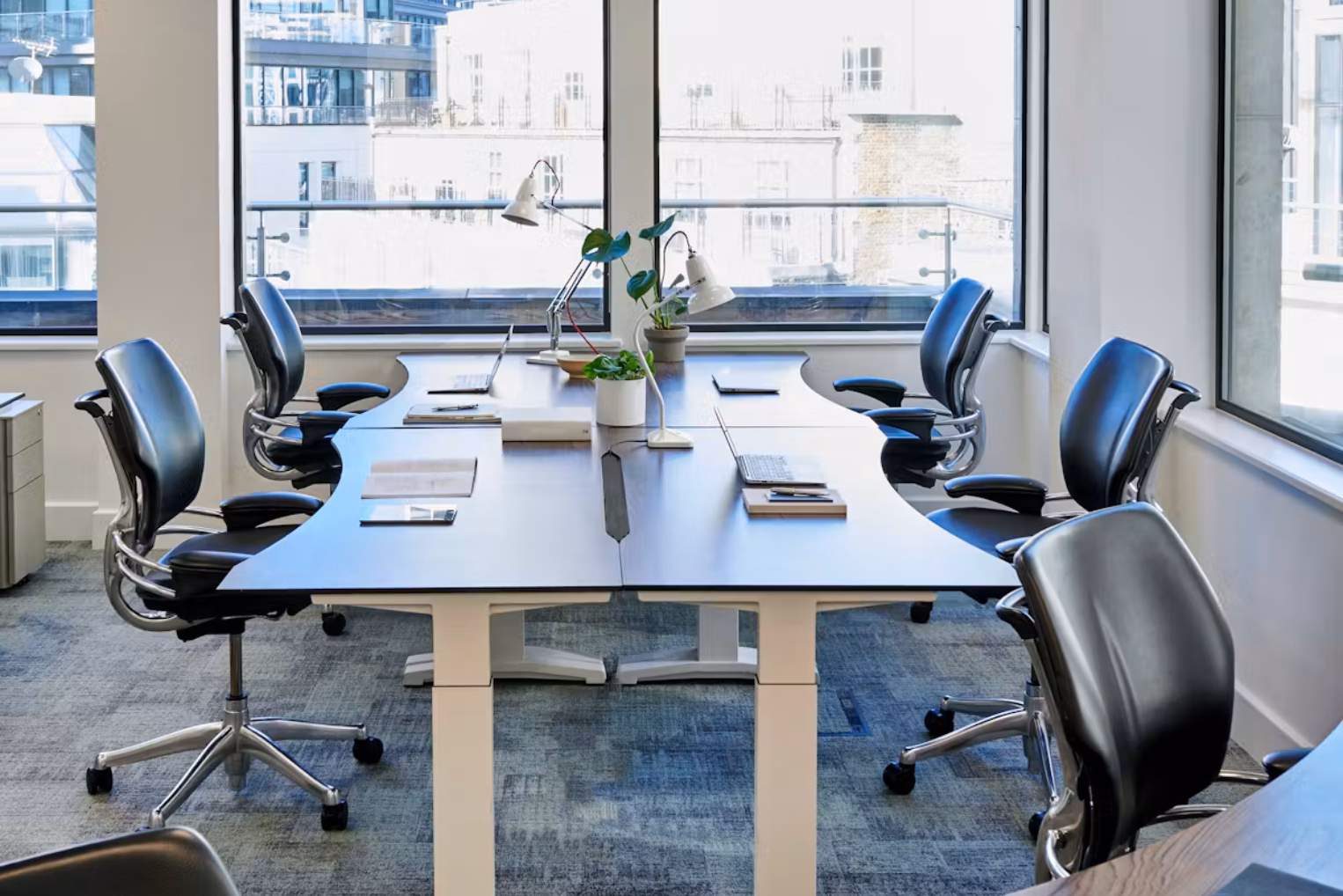 Black leather chairs at dark wood desks on a grey carpeted floor, with large windows looking out onto a city building.