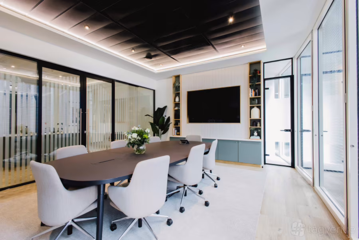Grey rolling chairs surround a brown table on a light-coloured rug in a boardroom with glass partition walls and a wall-mounted TV.