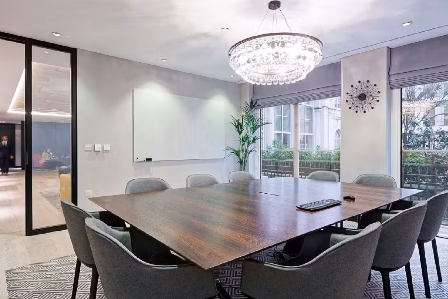 Grey chairs surround a dark wooden table on a geometric patterned rug, in a room with a whiteboard, a chandelier, and large windows.