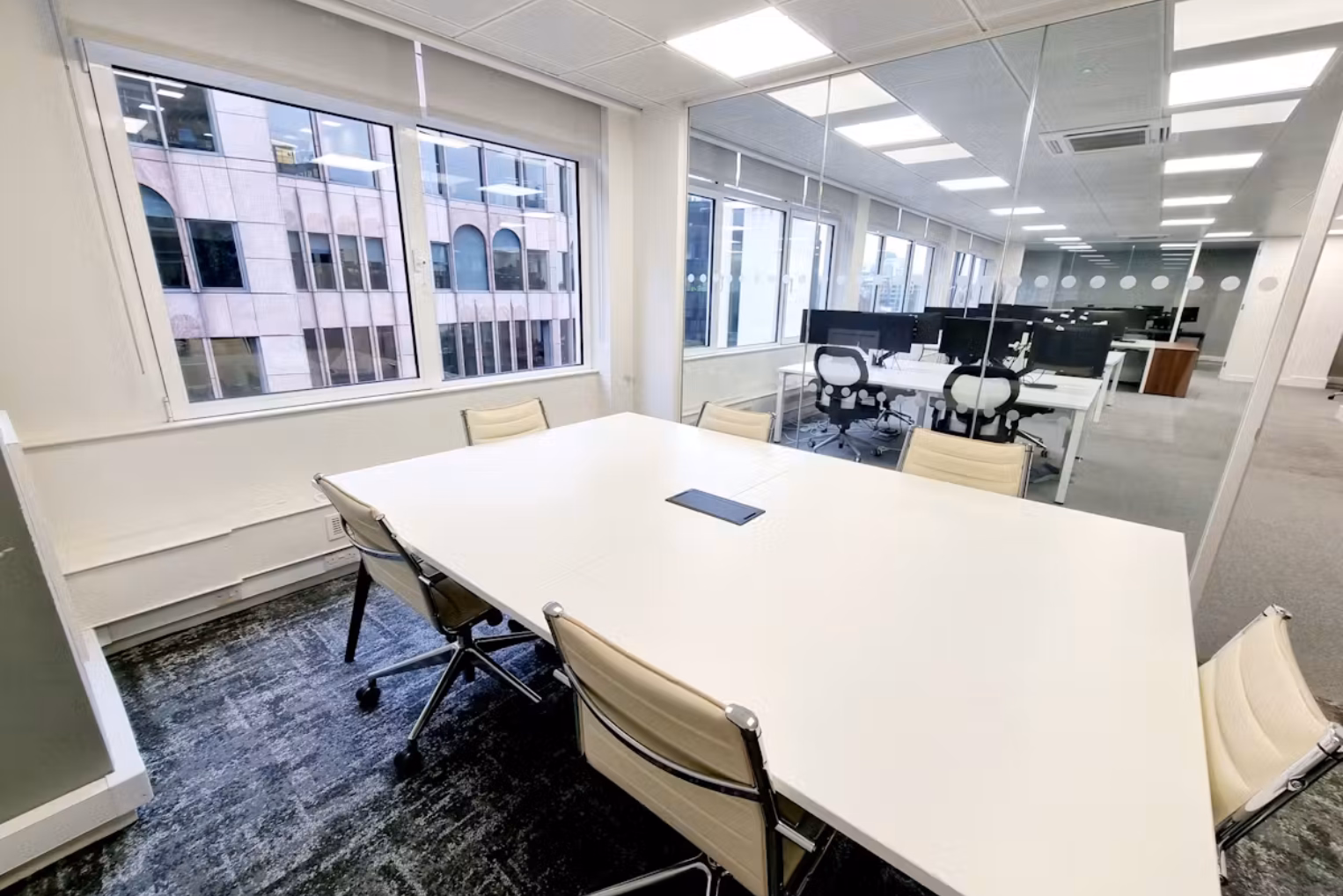 Cream-coloured chairs at a white table on a grey carpeted floor that features a glass wall looking into an open office space.