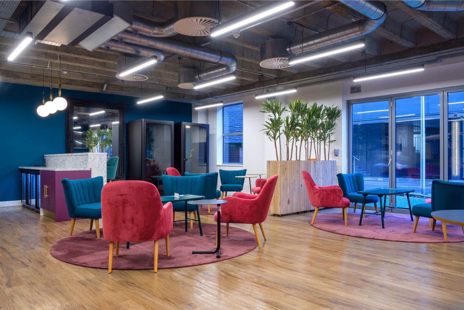 Magenta and teal armchairs around small tables on round rugs in a room featuring an industrial ceiling with exposed ductwork.