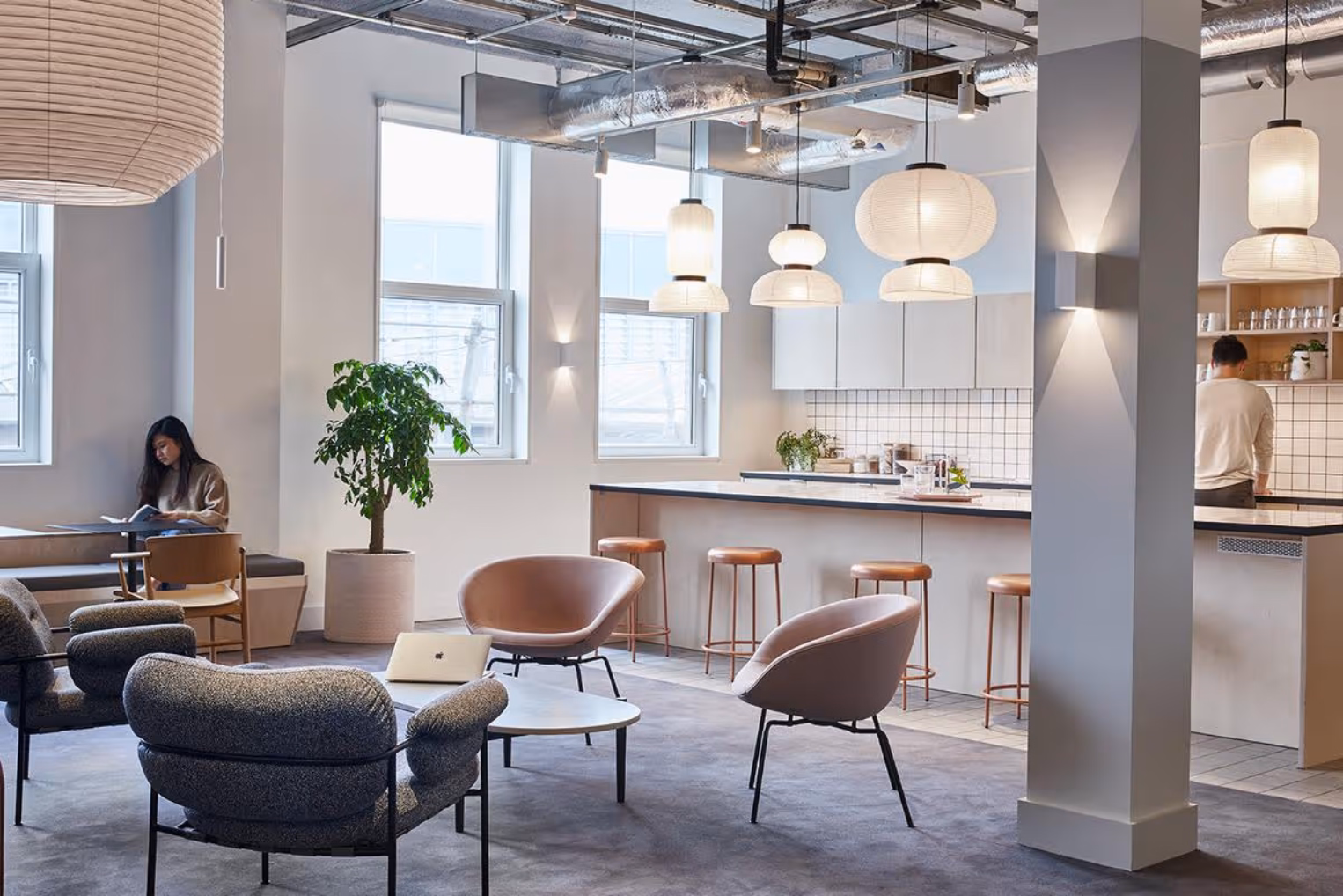 Grey armchairs and pink chairs around a low coffee table on a grey carpeted floor next to a kitchen bar area with light brown barstools and hanging white paper lantern lights.
