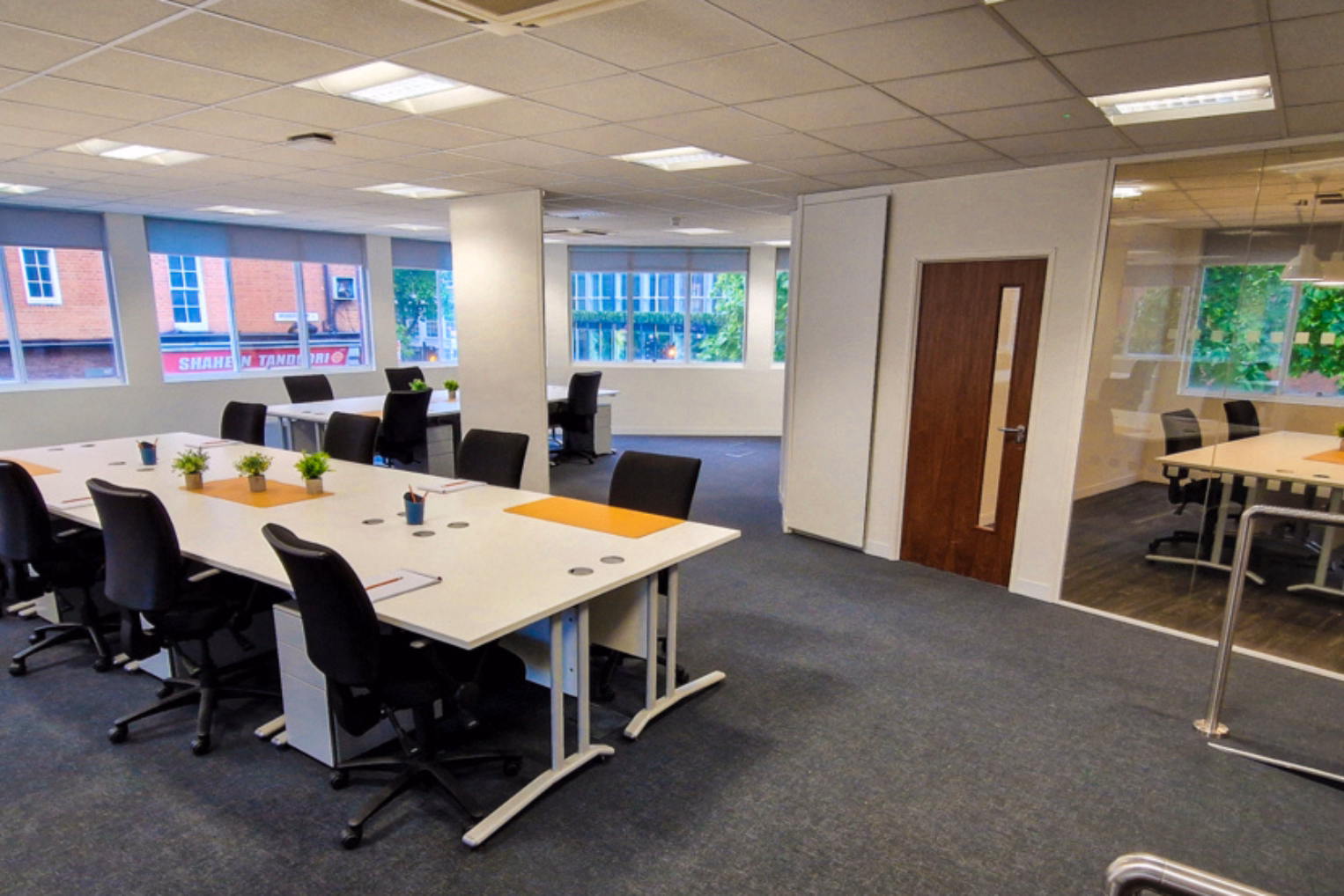 Black office chairs around white desks on a dark grey carpeted floor, in a bright office space with multiple windows.