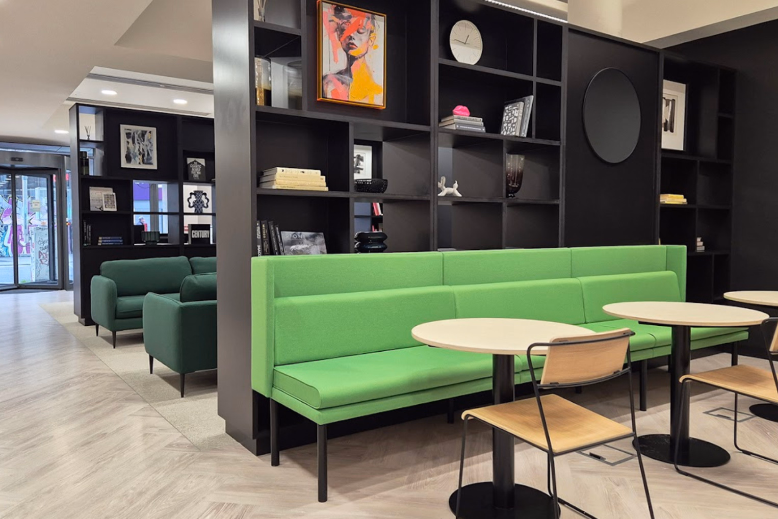 A bright green cushioned bench with round light-wood tables and chairs, in front of a grey open shelving unit.
