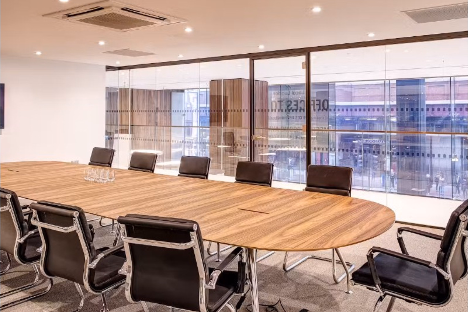 Black leather chairs surround a large oval wooden table on a grey carpeted floor in a room with a floor-to-ceiling glass partition wall.