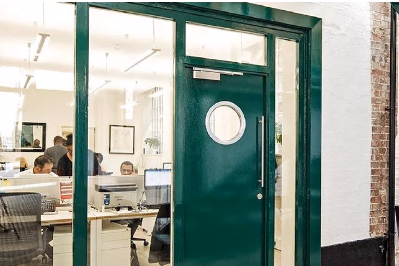 An office with employees working at white desks through a large glass partition and a green door with a round porthole window.