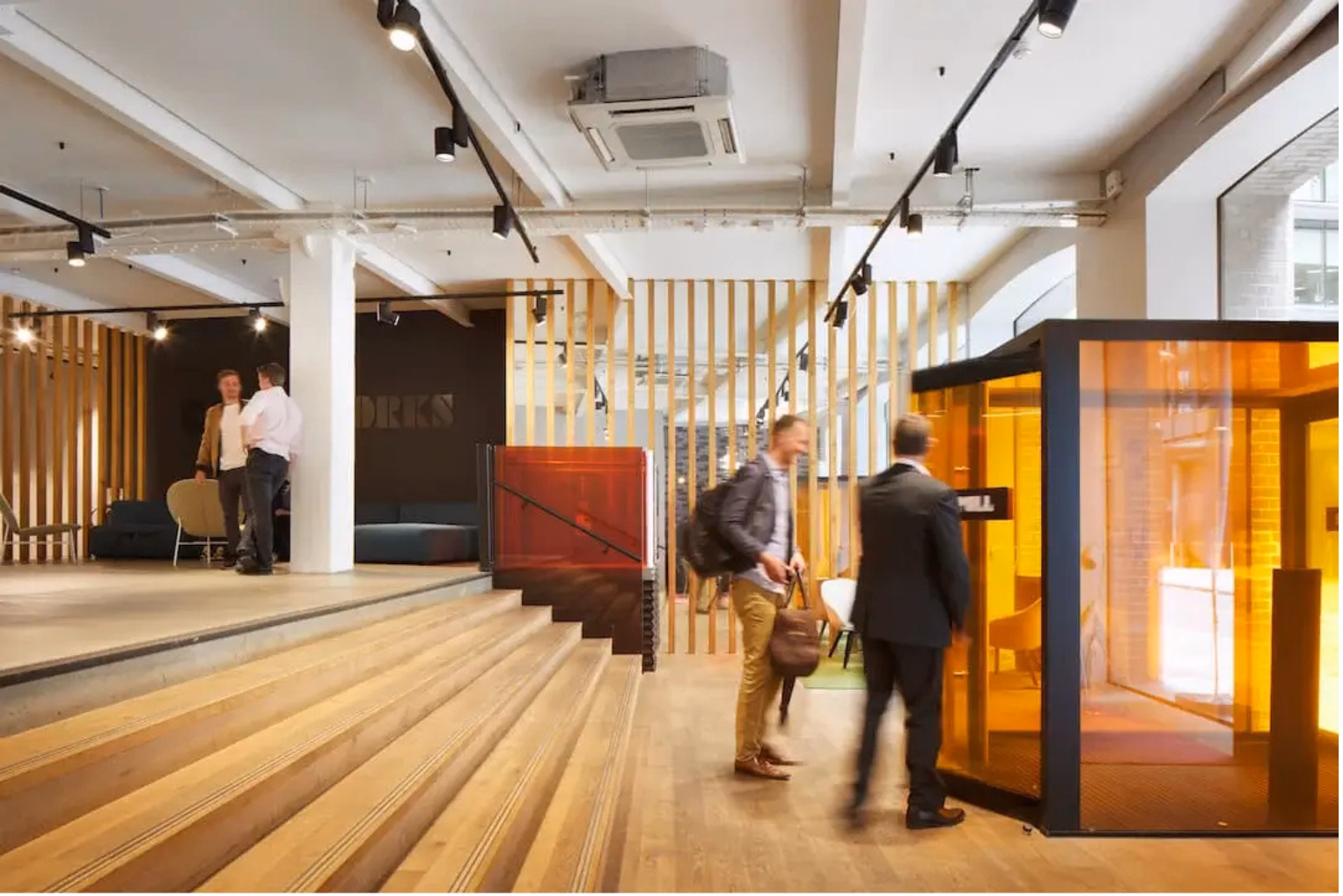 People stand and walk across light wood floors in an office lobby with track lighting, a wooden-slat partition, and an amber glass phone booth.
