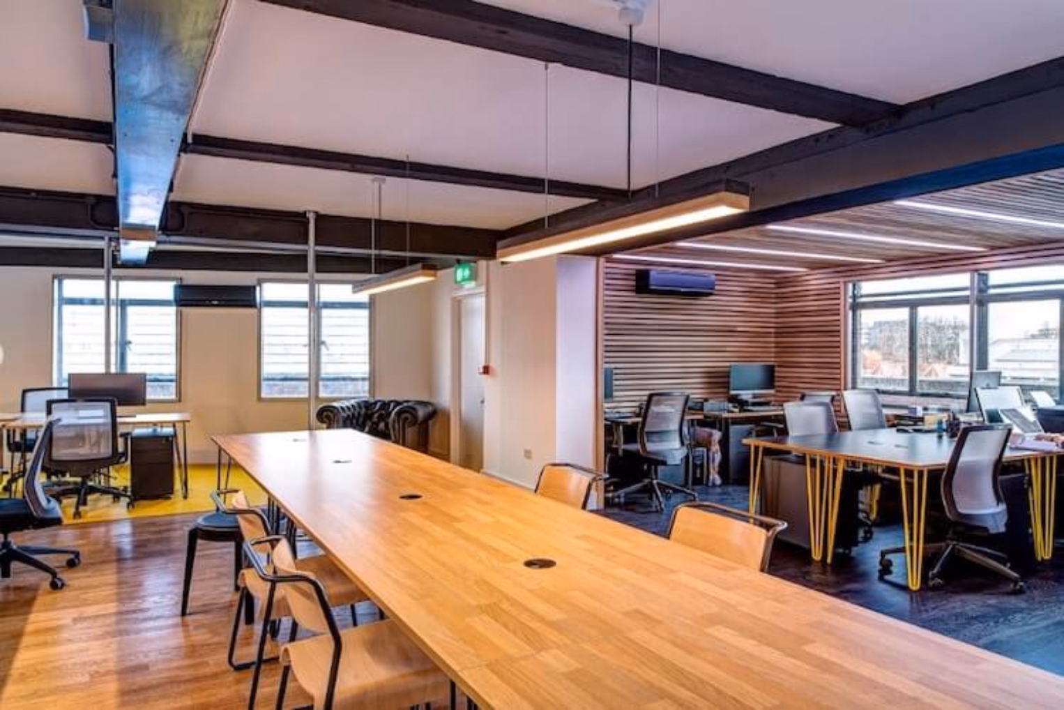 A long wooden table with light wood chairs and black mesh chairs around desks under exposed ceiling beams.