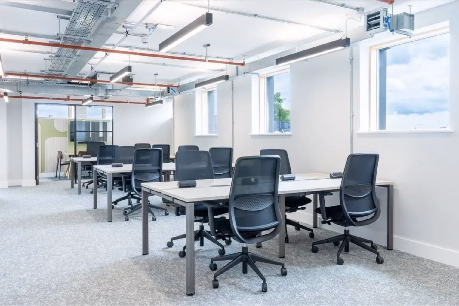 Black mesh chairs around white desks on a light grey carpeted floor, in a white-walled room with several windows and an industrial ceiling.